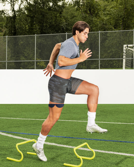 A man on a football field performing a jump drill, wearing a grey SAXX tee and black SAXX shorts. A horizontal section of the image is removed to reveal SAXX PRO performance underwear under his clothing.